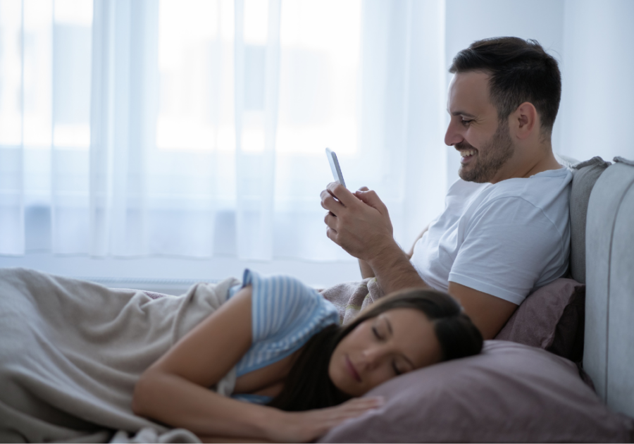man sitting in bed beside woman looking at phone Snapchat Cheating